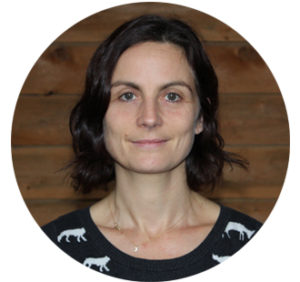 Portrait d'Élise, en couleur. Élise a la peau blanche et les cheveux bruns mi-longs. Elle pose devant un mur en bois et porte un chandail foncé avec des motifs d'animaux blancs. Elle sourit doucement en regardant la caméra.