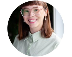 Portrait de Justine. Elle porte des lunettes en métal, des cheveux auburn aux épaules avec un toupet au carré. Elle sourit en regardant l'objectif.