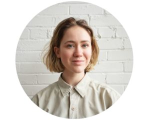 Portrait en couleur de Madeleine. Elle a les cheveux blonds légèrement frisés coupés au-dessus des épaules. Elle pose devant un mur de briques blanches et arbore un léger sourire.