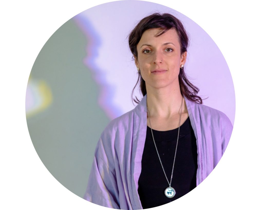 Portrait d'Ariane, en couleur. Elle a la peau blanche et les cheveux mi-longs chatains. Elle porte un chandail noir et une veste mauve lavande. Sur le mur blanc devant lequel elle pose, son ombre est visible en raison de l'éclairage.