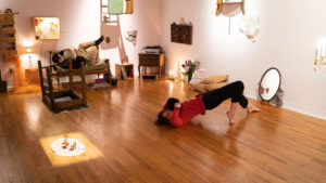 Dans une pièce aux murs blancs avec plancher de bois, avec du mobilier de salon disposé étrangement (une chaise avec les pattes clouées au mur, une petite table suspendue à l'envers du plafond, et d'autres meubles traditionnellement disposés, deux interprètes s'activent sans se regarder. L'un lit un livre, couché sur un divan, avec une jambe en l'air, l'autre a les épaules bien ancrées au col mais le dos et les genoux surélevés, les pieds pointés au sol.