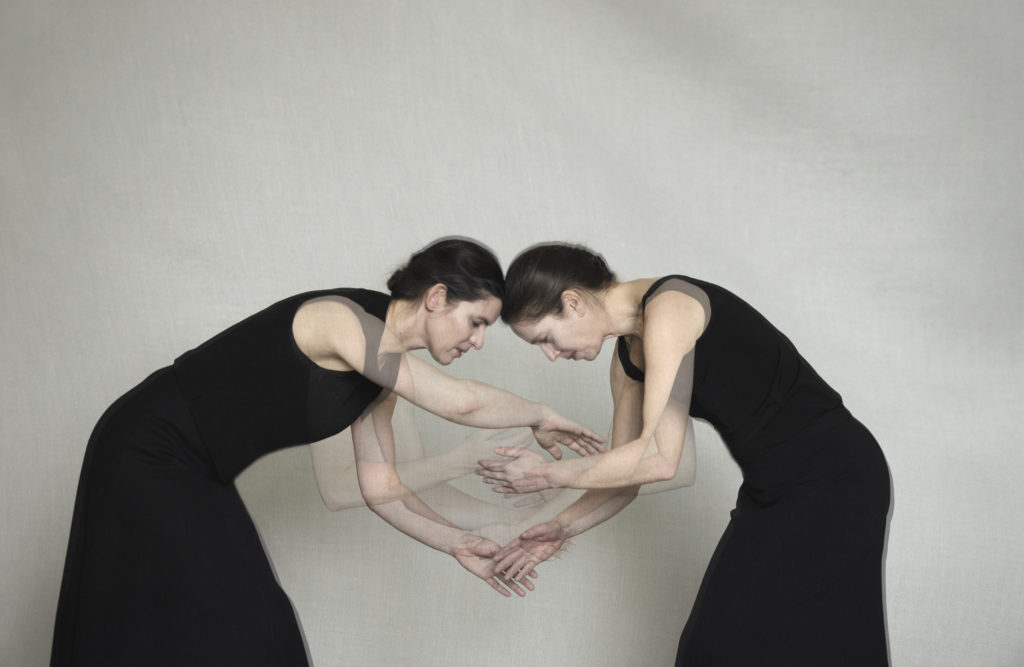 Deux femmes à la peau blanche, vêtues de robes noires, sont face à face, légèrement penchées l'une vers l'autres, leurs têtes collées au centre de l'image. Une superposition d'images en transparence montre leurs bras entremêlées entre elles deux.