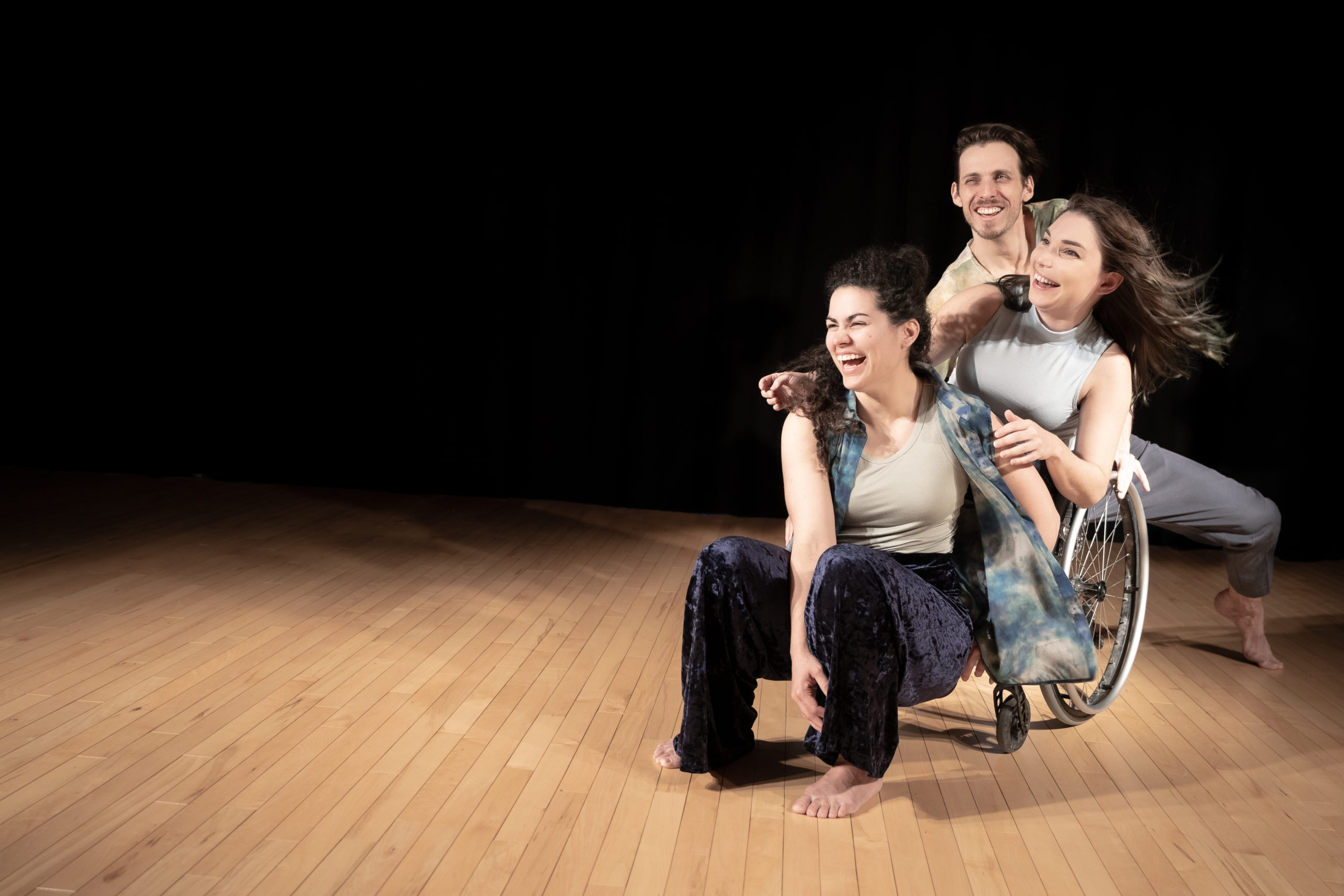 Les trois interprètes de Quand tu me regardes sont regroupés dans le côté droit de l'image, devant un rideau noir et sur un plancher de bois. Devant, Ivanie, cheveux foncés frisés attachés en demi-couette avec pantalons foncés et chemise à manche courte ouverte est assise sur l'appuie-pieds du fauteuil roulant de Maxime, cheveux lisses courts bruns, qui a les mains sur les épaules d'Ivanie. Derrière elles, Jérémie Desbiens, cheveux courts bruns et petite moustache est accroupi. On ne voit que sa tête et ses épaules. La photo est prise au moment d'un éclat de rire des trois interprètes qui montrent leur complicité.