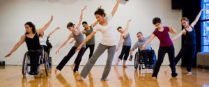 Une dizaine de personnes dansent dans un studio de danse avec plancher de bois. Certaines sont en fauteuil roulant, d'autres debout