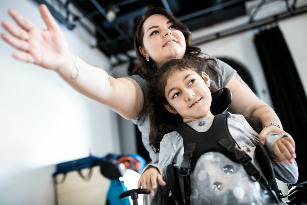 Ateliers : Une enfant en fauteuil motorisé regarde hors du cadre avec un air espiègle. Derrière elle, une accompagnatrice adulte tend le bras vers la caméra.