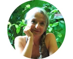 Portrait de Christine, en couleur. Elle a la peau blanche et les cheveux gris tressés. Elle regarde l'objectif en souriant, le visage appuyé sur son bras droit. Derrière elle, se trouve un fond végétal