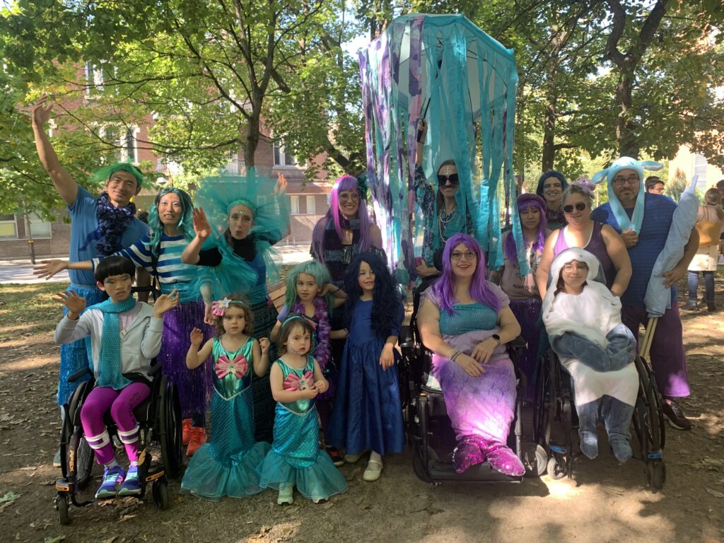 Un groupe de personnes costumées en sirènes, requins et créatures marines non-identifiées sourient à la caméra avant d'entrer dans la Parade phénoménale avec Corpuscule Danse.