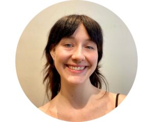 Portrait d'Aliénor, en couleur. Elle pose devant un mur blanc. Elle a la peau blanche, une coupe mulet, les cheveux bruns, un toupet séparé en deux au milieu. Elle fait un grand sourire à la caméra.