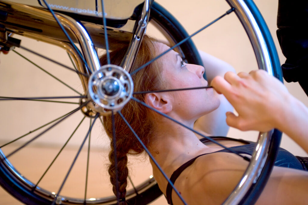 Une femme est couchée sous un fauteuil roulant dont elle tient les rayons des roues avec ses mains.