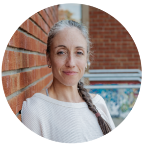 Portrait de Joannie en couleur. Devant un mur de briques rouges, elle est cadrée du haut de la tête à la poitrine. Elle porte un chandail blanc. Ses cheveux sont assemblés en une tresse qui tombe sur son épaule gauche. Elle a les yeux clairs et un visage serein.