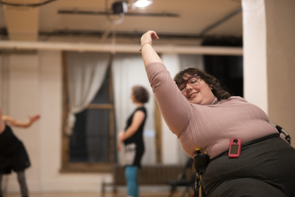 Une photo en couleur présente une femme au chandail rose sur la gauche de la photo. Elle danse en levant le bras, et en tournant le visage vers son épaule, dans un fauteuil roulant. Derrière elle, deux autres personnes dansent dans la salle de danse devant des rideaux blancs.