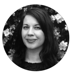 Portrait de Sarah, en noir et blanc. Elle a la peau blanche et les cheveux foncés. Elle pose devant un arbre en fleurs contrasté. Elle porte un chandail en dentelle foncé sans manches. Son visage est de 3/4 et elle sourit légèrement en regardant la caméra.