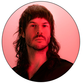 Portrait de Simon, en couleur. Sa peau blanche est éclairée par une lumière rouge. Il porte une coupe mulet avec un toupet et une petite barbe. Ses cheveux sont bruns. Il fixe l'objectif d'un air sérieux et fier.