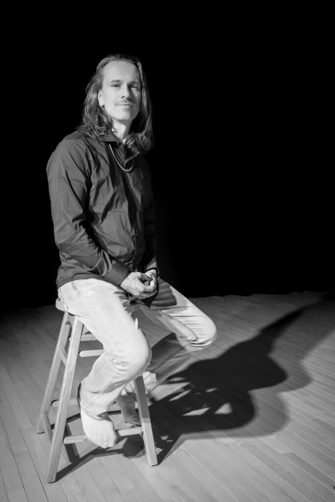 Georges-Nicolas est assis sur un tabouret qui laisse voir sa silhouette de sa tête à ses pieds. Il porte une chemise foncée et un pantalon beige. Il est tourné de 3/4 vers la droite de la photo. Il a la peau claire, porte des cheveux longs lâchés et arbore un léger sourire.